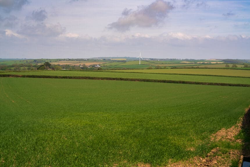 Green farm fields under a flat overcast sky with a distant wind turbine on the horizon
