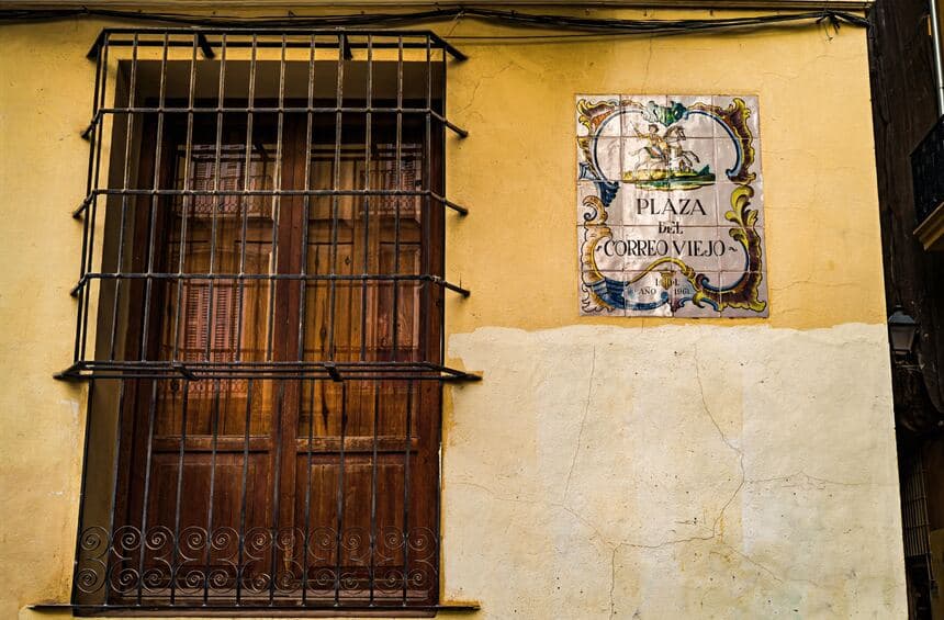 Ochre Spanish wall with a barred window and a painted ceramic tile sign reading Plaza del Correo Viejo